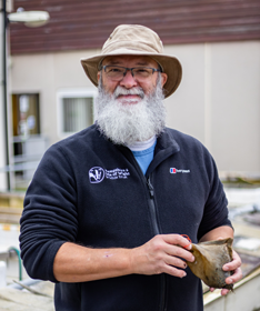 Image shows Dr Tim Ferrero, who is a white bearded man with a friendly expression. He is wearing silver-coloured eyeglasses and a beige bucket-style sunhat. Dr Ferrero is holding what appears to be a shark or ray egg case.